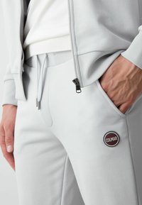 Gaiši pelēki sporta bikses, izgatavotas no kokvilnas maisījuma, ar rāvējslēdzēja kabatu un apaļu logo uzlīmi kreisajā augšstilbā. Mīksta tekstūra, pieguļošs dizains.