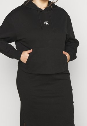 Woman wearing black Calvin Klein hoodie with front pocket and matching black skirt, hands in pockets, standing against plain background.