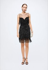 Black sequined dress with a sheer neckline, textured fringe hem, fitted bodice, and thin spaghetti straps, paired with strappy black heels.
