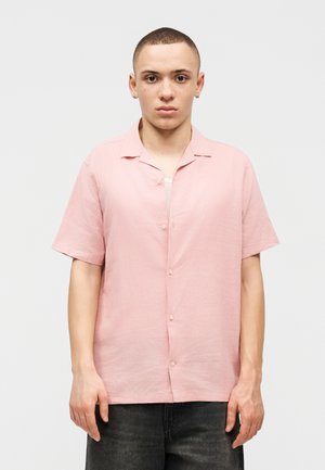 Jeune personne aux cheveux courts portant une chemise à manches courtes rose clair boutonnée et un pantalon foncé, debout devant un fond blanc uni.