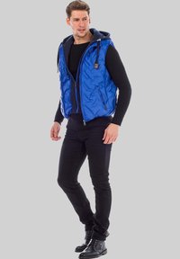 Blau gesteppte Weste mit Reißverschluss und Kapuze, getragen über einem schwarzen strukturierten Pullover, kombiniert mit schwarzen Hosen und schwarzen Schnürstiefeln.