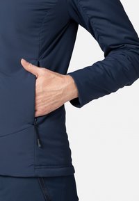 Marineblaue Softshelljacke mit einer Reißverschlusstasche an der unteren Seite, aus glattem Material und mit tailliertem Schnitt.