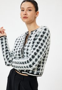 Teksturoitu musta-valkoinen lyhytnen cardigan, jossa on houndstooth-kuvio, ribbireunat mustana ja valkoisena sekä etupainike.