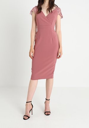 Femme portant une robe rose poudrée ajustée avec des manches courtes en dentelle et des sandales noires à talons hauts avec bride à la cheville, debout sur un fond blanc.