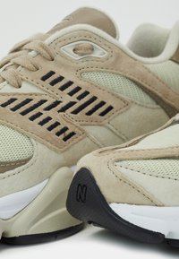 Gros plan sur des baskets beige et vert clair avec des détails noirs cousus et une semelle noire texturée avec logo en relief.