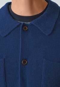 Granatowy sweter dzianinowy z wyostrzonym kołnierzem, dwoma przednimi kieszeniami i brązowymi guzikami. Teksturowana tkanina o miękkim wyglądzie.