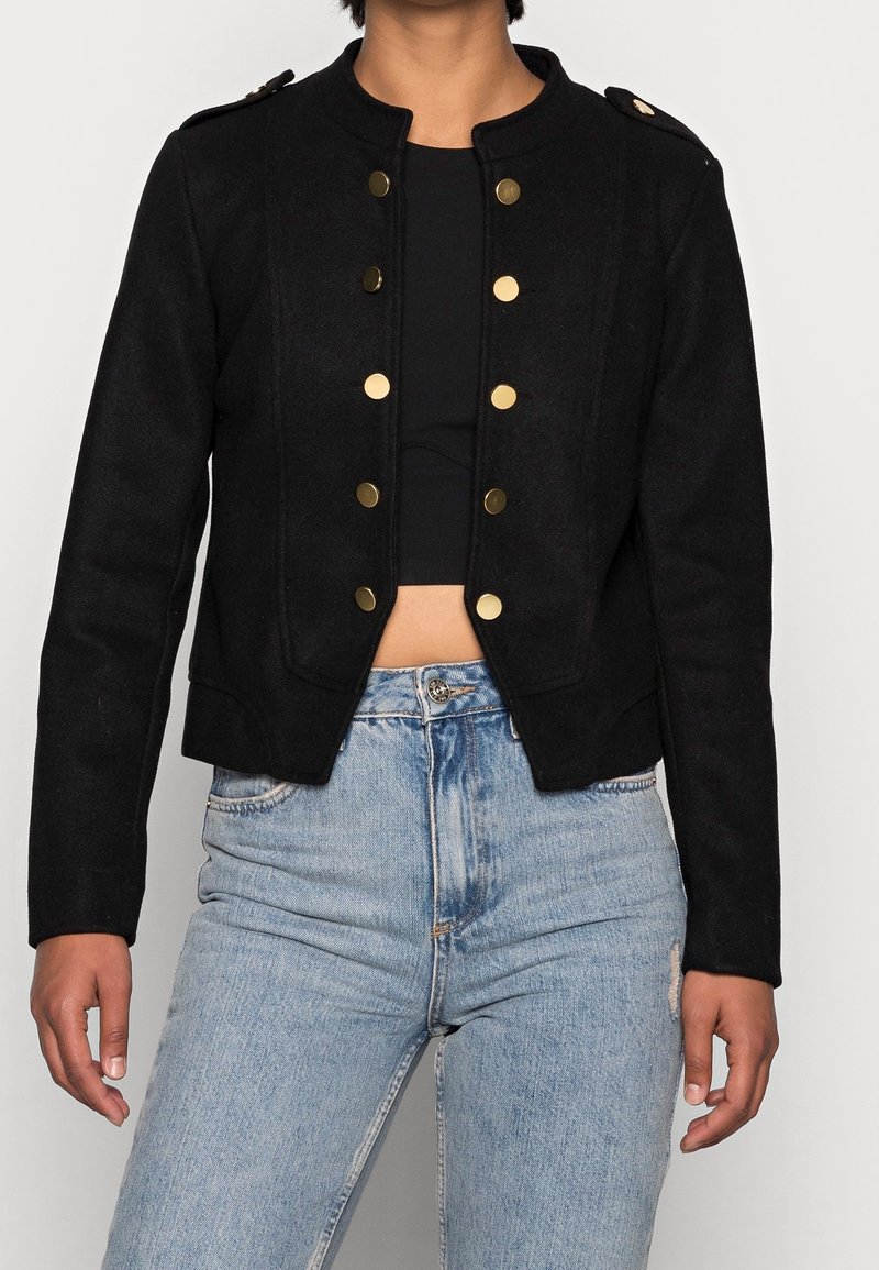 Svart kort jacka med guldknappar, strukturerad design och rund krage, kombinerad med högmidjade ljusa jeans med slitningar.