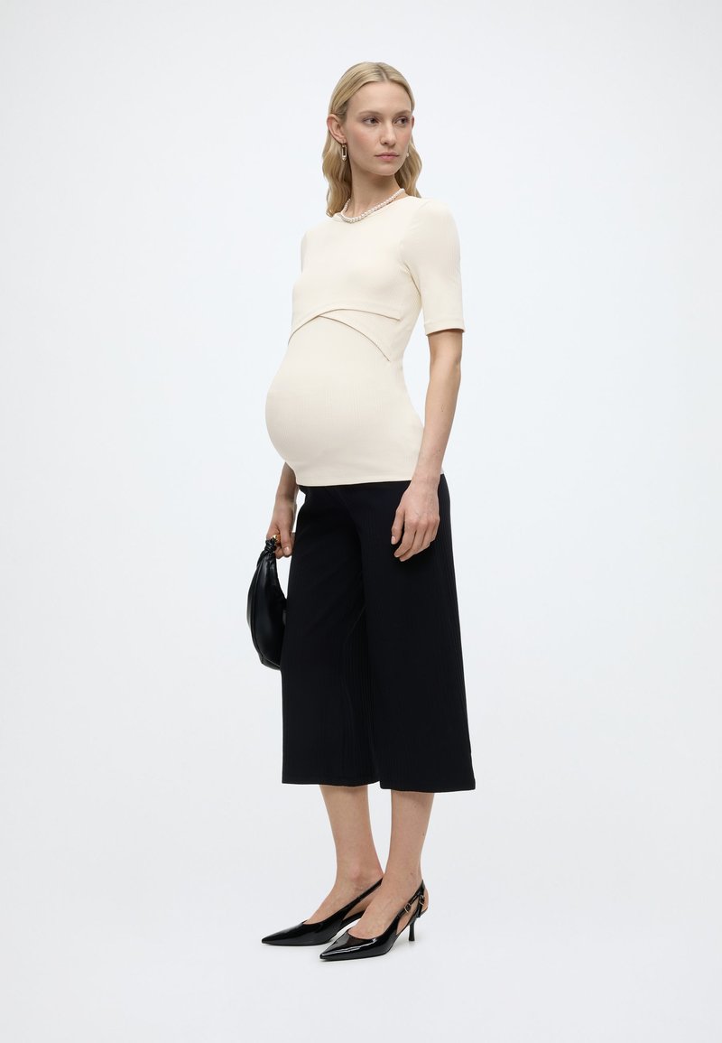 Έγκυος γυναίκα που φοράει ένα εφαρμοστό κρεμ τοπ, μαύρο φαρδύ παντελόνι cropped, μαύρα τακούνια, κρατώντας μια μαύρη τσάντα, στέκεται μπροστά σε λευκό φόντο.