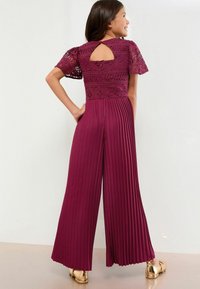 Salopetă burgundy cu un corp din dantelă cu modele geometrice, mâneci scurte, picioare ample pliate și un detaliu cu deschidere în formă de cheie la spate.