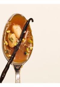 Cuillère avec sauce caramel, amandes entières, pistaches hachées, flocons de sel marin et une gousse de vanille entière sur un fond clair.