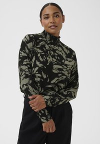 Langarmshirt in zwart met een olijfgroen bladpatroon. Heeft een hoge, gerimpelde halslijn en een gestructureerde stof. Gecombineerd met zwarte broek.