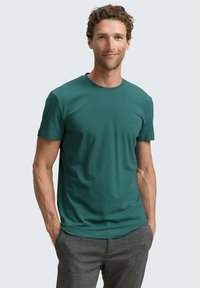 Turkusowy t-shirt z okrągłym dekoltem, wykonany z miękkiego materiału, z krótkimi rękawami. Dopasowany krój podkreśla kształt górnej części ciała; noszony z szarymi spodniami.