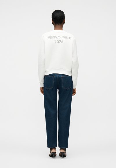 Γυναίκα με σκουρόχρωμο δέρμα και μαλλιά σε κότσο, φορούσε λευκό φούτερ με την επιγραφή «SPRING/SUMMER 2026», σκούρο μπλε τζιν και μαύρα πέδιλα με ψηλό τακούνι.