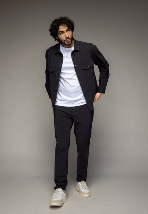 Zwarte jas met twee borstzakken over een wit T-shirt, gecombineerd met zwarte broek en lichtgrijze sneakers. Glad materiaal, casual pasvorm.