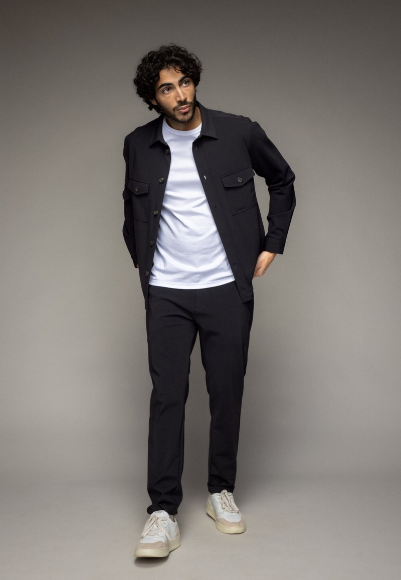 Black jacket with two chest pockets over a white t-shirt, paired with black pants and light gray sneakers. Smooth fabric, casual fit.