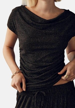 Femme portant un top noir scintillant à manches courtes et un pantalon assorti, ajustant la taille, avec un bracelet en or et une bague.