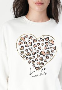 Weißes Sweatshirt mit einem großen, herzförmigen Muster aus pinken und schwarzen Herzen, umrandet von Gold, sowie dem Text "Liebe versagt nie" darunter.