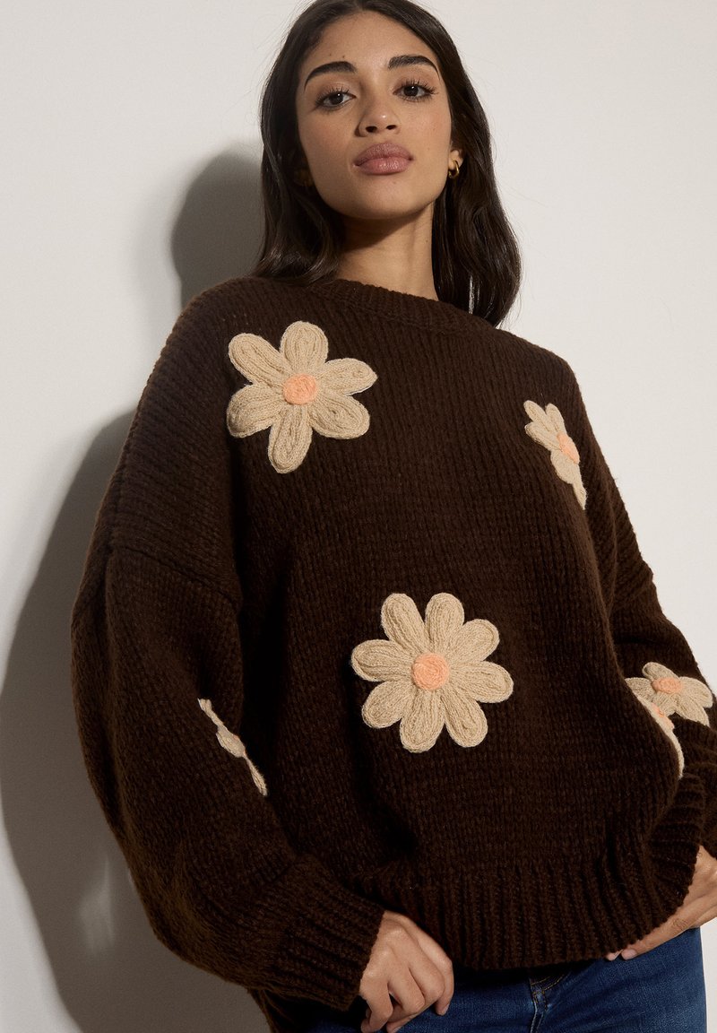 Femme portant un pull en tricot marron avec de grands motifs floraux crème et pêche, debout devant un fond clair uni.