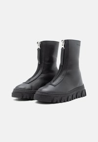 Bottes noires en cuir à la cheville avec des zippers argentés, semelles en caoutchouc texturées et épaisses, et un design épuré et minimaliste. Présente des détails de couture visibles.