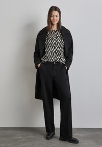 Czarny płaszcz na wzorzystej czarno-kremowej bluzce w zygzakowy wzór; zestawiony z czarnymi spodniami o szerokich nogawkach i czarnymi butami. Gładki, dopasowany materiał.