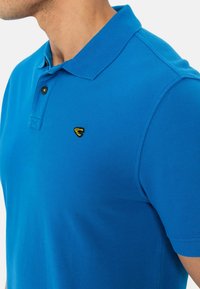 Niebieska koszulka polo z teksturowanej tkaniny, z klasycznym kołnierzykiem, zapięciem na dwa guziki i małym żółtym logo na lewej piersi.