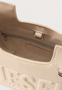 Μπεζ υφασμάτινη τσάντα χειρός με εμφανές εσωτερικό φερμουάρ στην τσέπη και ανάγλυφο όνομα της μάρκας στην εξωτερική μπροστινή πλευρά.