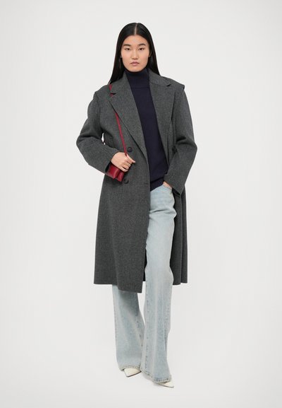 Cappotto di lana grigio con grandi rever e doppi bottoni, abbinato a un dolcevita blu navy e jeans a zampa d'elefante azzurri. Borsa a tracolla rossa.
