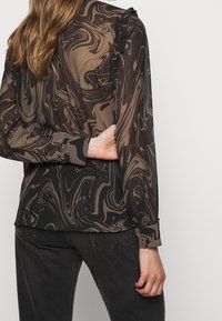Vrouw in een blouse met lange mouwen, zwart en bruin met een spiraalpatroon en ruches, gecombineerd met zwarte denimjeans, hand achter haar rug.