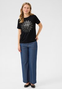 Sort t-shirt med et sølvfarvet mandala-mønster, parret med vide blå denim jeans med kontrastsyninger. Sorte flade sko.