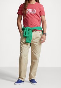 Röd bomulls t-shirt med "Polo Ralph Lauren"-logotyp, khakifärgade byxor, grön stickad tröja knuten runt midjan och blå sneakers. Silverfärgad klocka på handleden.