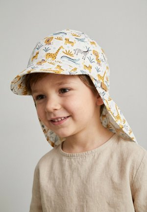 Junge lächelt, trägt ein beiges Hemd und einen weißen Sonnenhut mit Tiermotiven, darunter Löwen, Giraffen, Zebras und Krokodile.