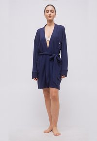 Γυναίκα που στέκεται ξυπόλητη, φοράει μπλε ναυτικό ρόμπα με δεμένη ζώνη και λευκό σουτιέν από κάτω, μπροστά σε απλό ανοιχτό φόντο.