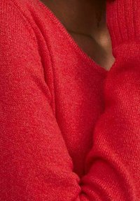 Pull rouge en maille avec un col en V. Présente une texture douce et des poignets côtelés, mettant en valeur un gros plan sur le tissu et les détails de couleur.
