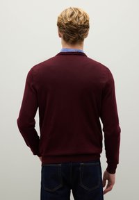 Cardigan a maglia bordeaux con polsini e orlo a costine, che mostra una superficie testurizzata. Il modello indossa una camicia con colletto sotto.