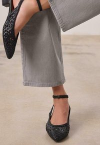 Chaussures plates tissées noires à bout pointu et bride à la cheville, avec un design texturé et des découpes, associées à un pantalon gris à jambes larges.