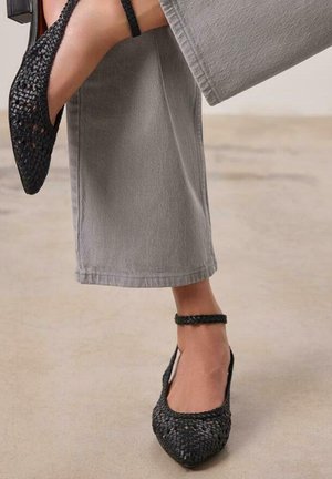 Zapatos planos negros de tejido trenzado con punta afilada y correa en el tobillo, con un diseño texturizado y detalles recortados, combinados con pantalones de pierna ancha grises.