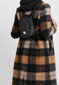 Svart ryggsäck med geometriskt mönster och justerbara remmar, med en framficka med dragkedja och vitt Adidas-logotyp, bärs över en rutigt kappa.