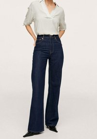 Nainen, jolla on päällään kääritaskuilla varustettu valkoinen pusero, korkeavyötäröiset tummansiniset leveälahkeiset farkut ja mustat kärkitossut, kädet selän takana.