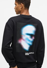 Must mustreeritud must فونь, mis sisaldab tekstuuriga graafikat sinise, roosa ja valge tooni näost. Brändi logo 'KARL LAGERFELD JEANS' valges.