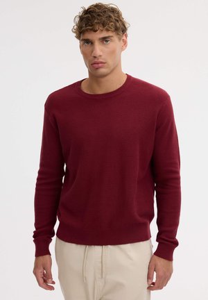 Un modelo masculino está de pie sobre un fondo claro, con un jersey burdeos de cuello redondo y textura marcada y unos pantalones color crema.