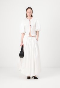 Blusa bianca a maniche a sbuffo abbinata a una gonna bianca a tutta lunghezza. Il modello tiene in mano una borsa nera e indossa tacchi a punta neri.