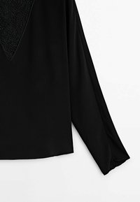 Vêtement noir à manches longues avec un panneau triangulaire texturé comportant de petits détails en perles près de l'épaule.