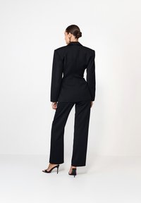 Blazer negru croit cu umeri pronunțați, asociat cu pantaloni negri drept și tocuri stiletto negre, văzut din spate.