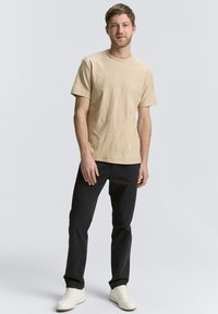 Tricou din bumbac bej cu un model subtil de palmier, asortat cu pantaloni negri și adidași albi, stând în fața unui fundal neutru.