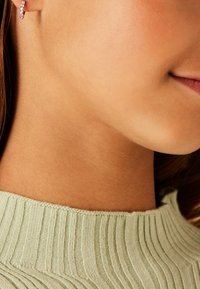 Zilveren hoepeloorbel met kleine doorzichtige kristallen, gecombineerd met een lichtgroene geribbelde trui. Close-up van de nek en onderkant van het gezicht.