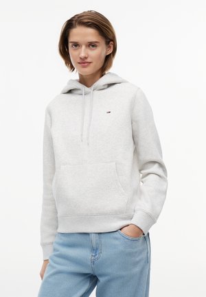 Νεαρή γυναίκα που φοράει ανοιχτό γκρι φούτερ με κουκούλα και μπροστινή τσέπη, διακοσμημένο με μικρό λογότυπο, συνδυασμένο με ανοιχτό μπλε τζιν, όρθια με το χέρι στην τσέπη.