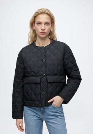 Junge Frau mit blonden Haaren, die eine schwarze gesteppte Jacke mit Knöpfen und blaue Jeans trägt, steht vor einem einfarbigen hellen Hintergrund.