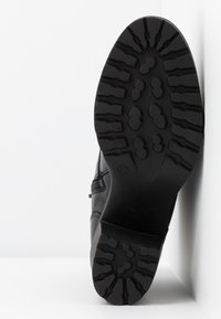 Semelle de chaussure noire avec motif de bande de roulement texturé et accents en relief. Dispose d'un talon incurvé et d'un logo. Présentée en angle sur un fond blanc.