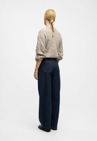 Frau mit blonden Zöpfen, die einen beigen Strickpullover, eine dunkle weite Jeans und schwarze Schuhe trägt, steht mit dem Rücken zu einer schlichten weißen Hintergrund.