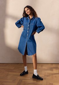 Denim-paitamekko vaaleansinisena, jossa on suuret hihat, nappilista, kaksi rintataskua ja korkeampi takakappale. Mustat kengät ja valkoiset sukat.